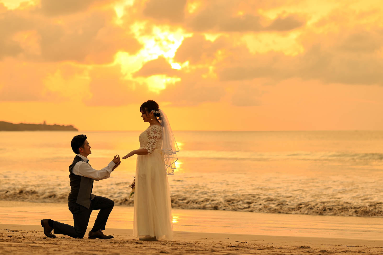 夕暮れのビーチでプロポーズシーンを再現する新郎新婦の写真
